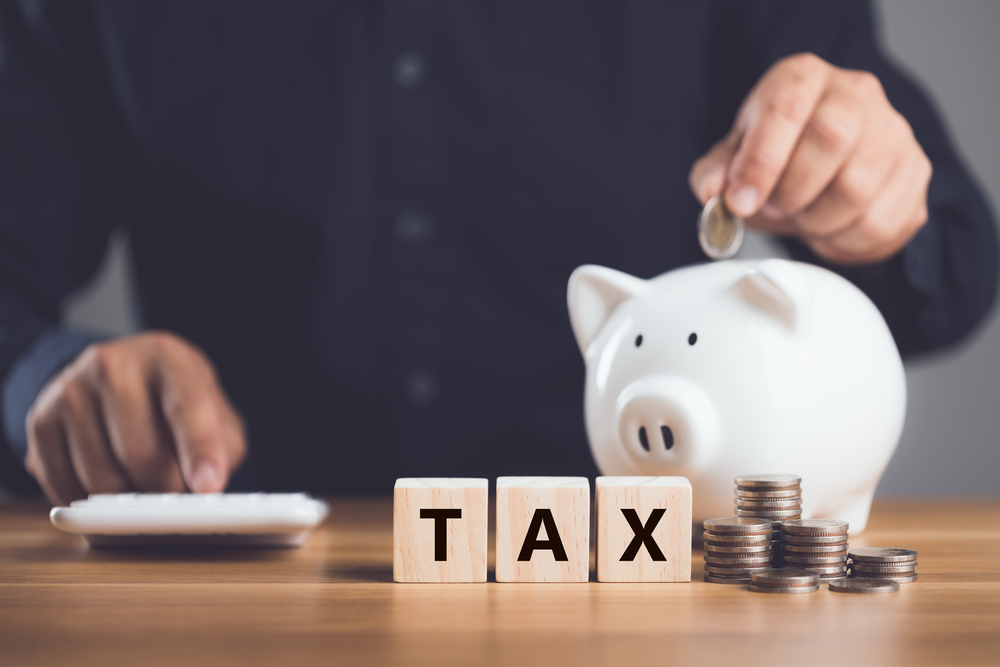 Man with calculator placing coins in piggy bank next to wooden blocks reading TAX