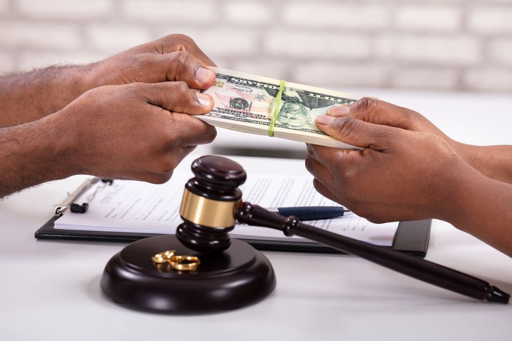 Two people fighting over stack of money next to gavel and wedding rings