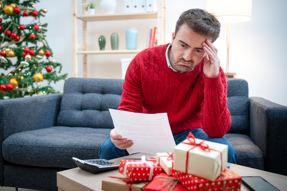 Upset man looking at paper with calculator and Christmas presents on coffee table