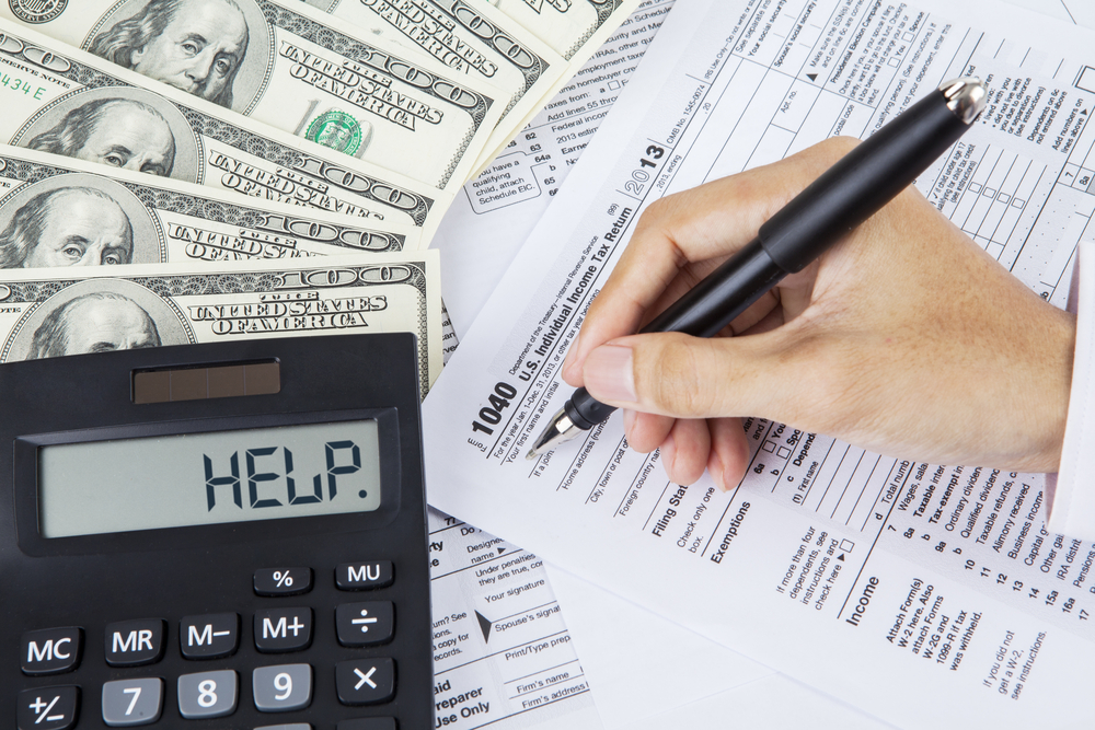 Hand filling out tax form near $100 bills and calculator that reads HELP