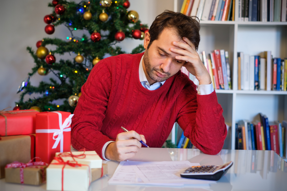 Worried man looking at paper and calculator with Christmas presents and tree nearby