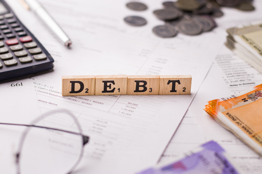 Debt on wooden blocks atop desk cluttered with coins, calculator, and other items