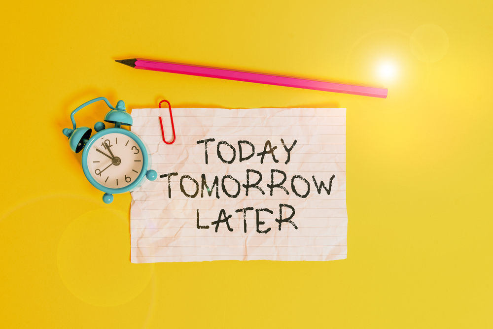 "Today Tomorrow Later" written on paper next to clock and pink pencil on yellow backdrop